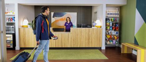 A hotel reception area with a wooden reception desk, a receptionist, a luggage trolley, and a green carpet.