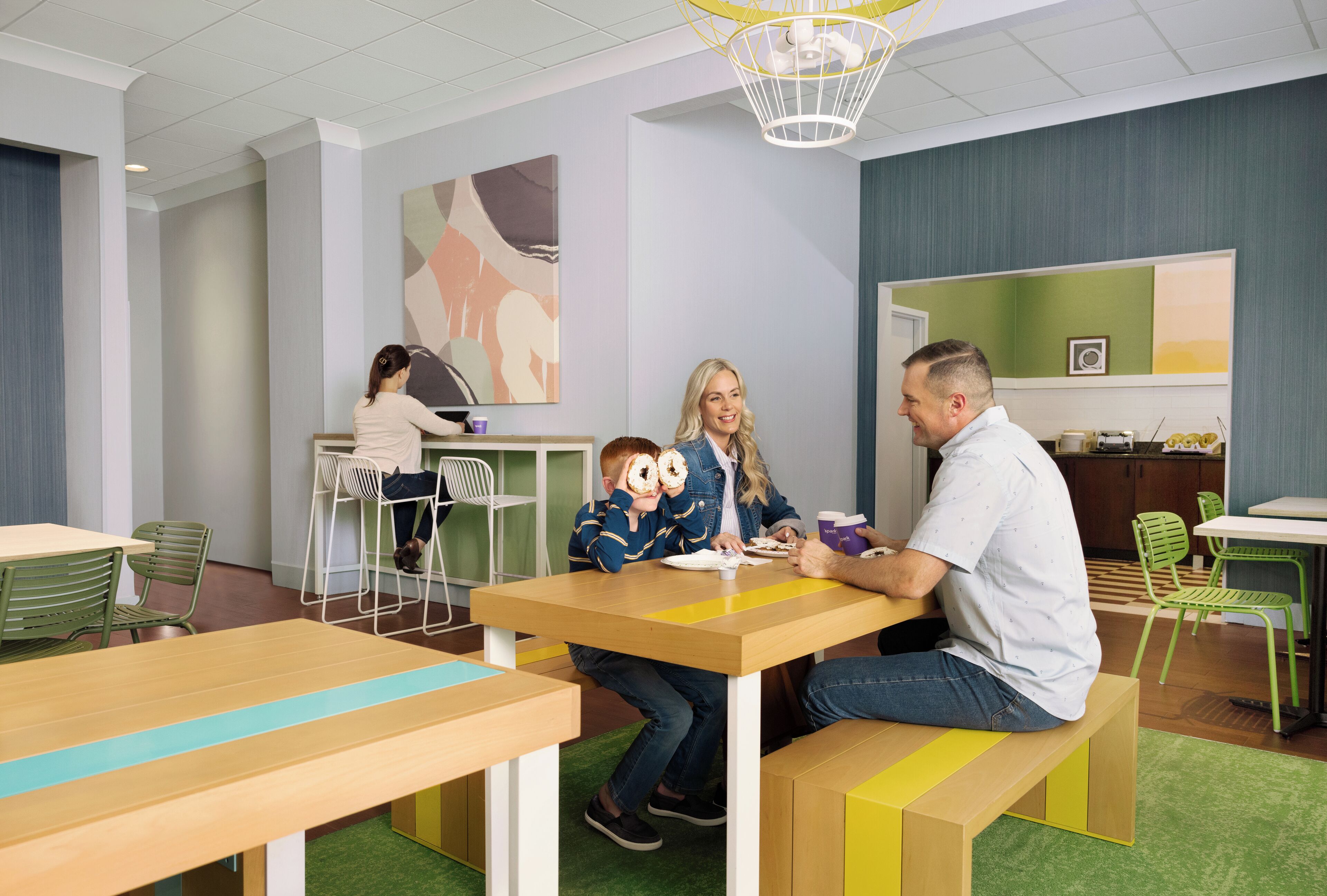 Een moderne binnenruimte met houten tafels en banken, groene stoelen en een groot abstract schilderij aan de wand.