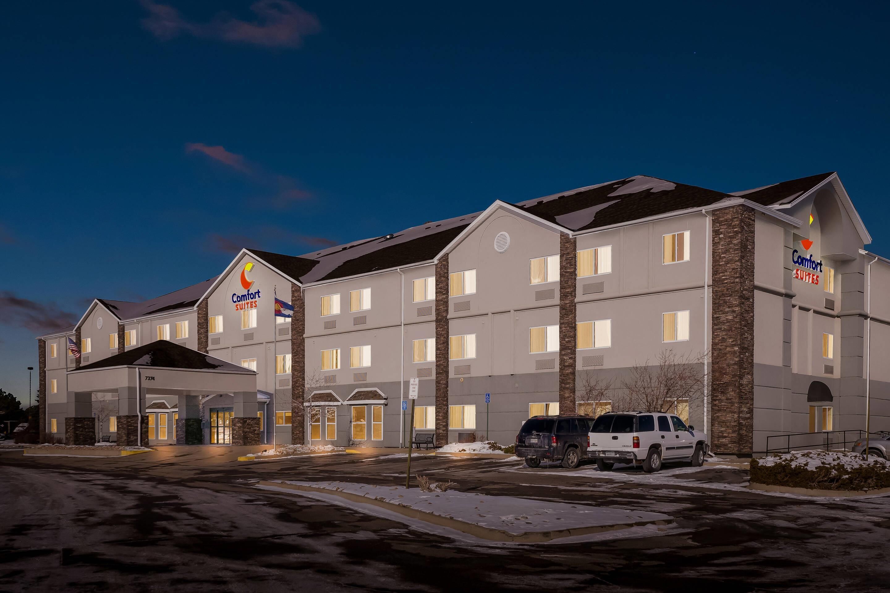 A multi-story hotel with a central entrance, multiple windows, and a parking area with several vehicles.