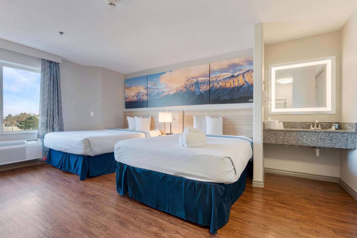 A hotel room with two beds, a large mountain landscape painting, a bathroom with a vanity and mirror, and a window with curtains.
