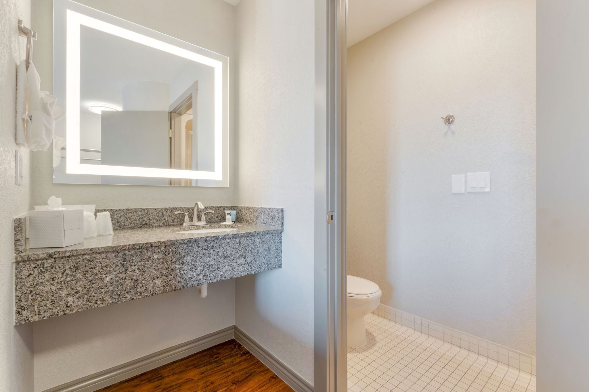 A bathroom with a granite countertop sink, a mirror with integrated lighting, a toilet, and a tiled floor.