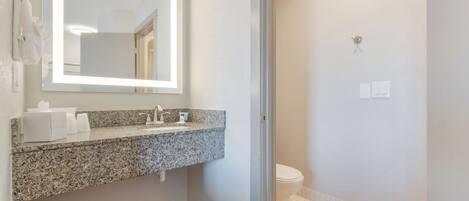 A bathroom with a granite countertop sink, a mirror with integrated lighting, a toilet, and a tiled floor.