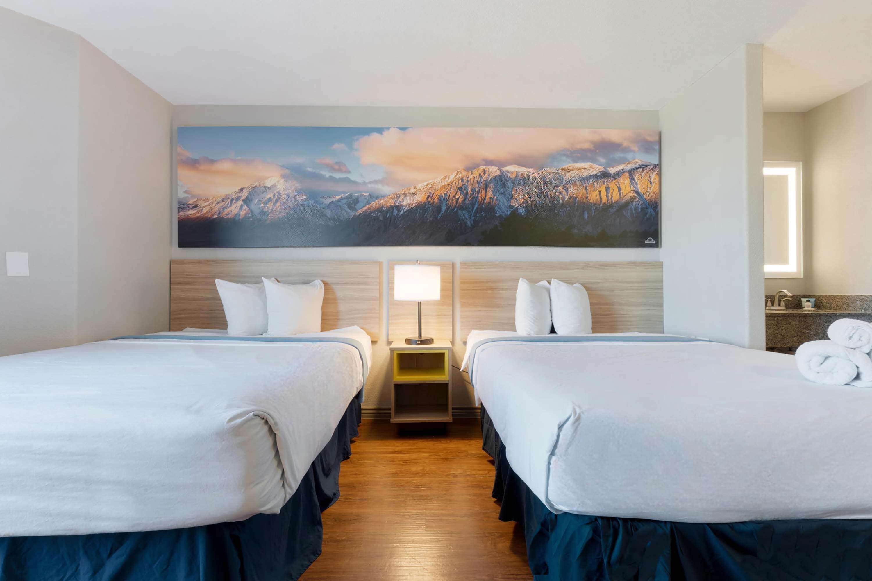 A hotel room with two beds, a large mountain painting on the wall, a bedside table with a lamp, and a view of the mountains.