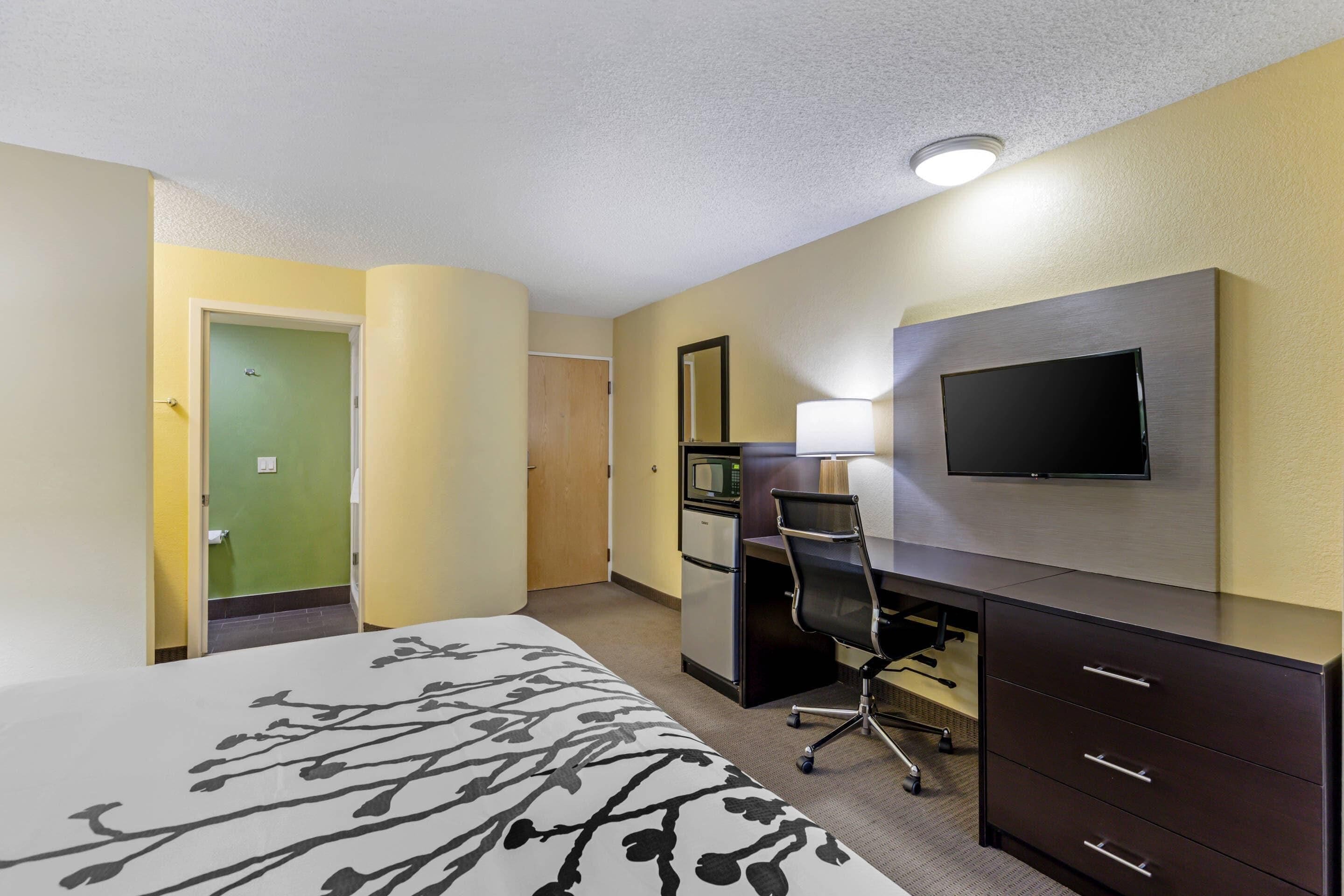 A hotel room with a bed, desk, chair, TV, and a small kitchenette.