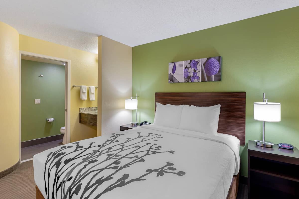 A hotel room with a bed, nightstands, a wall-mounted lamp, a painting, and a bathroom door.