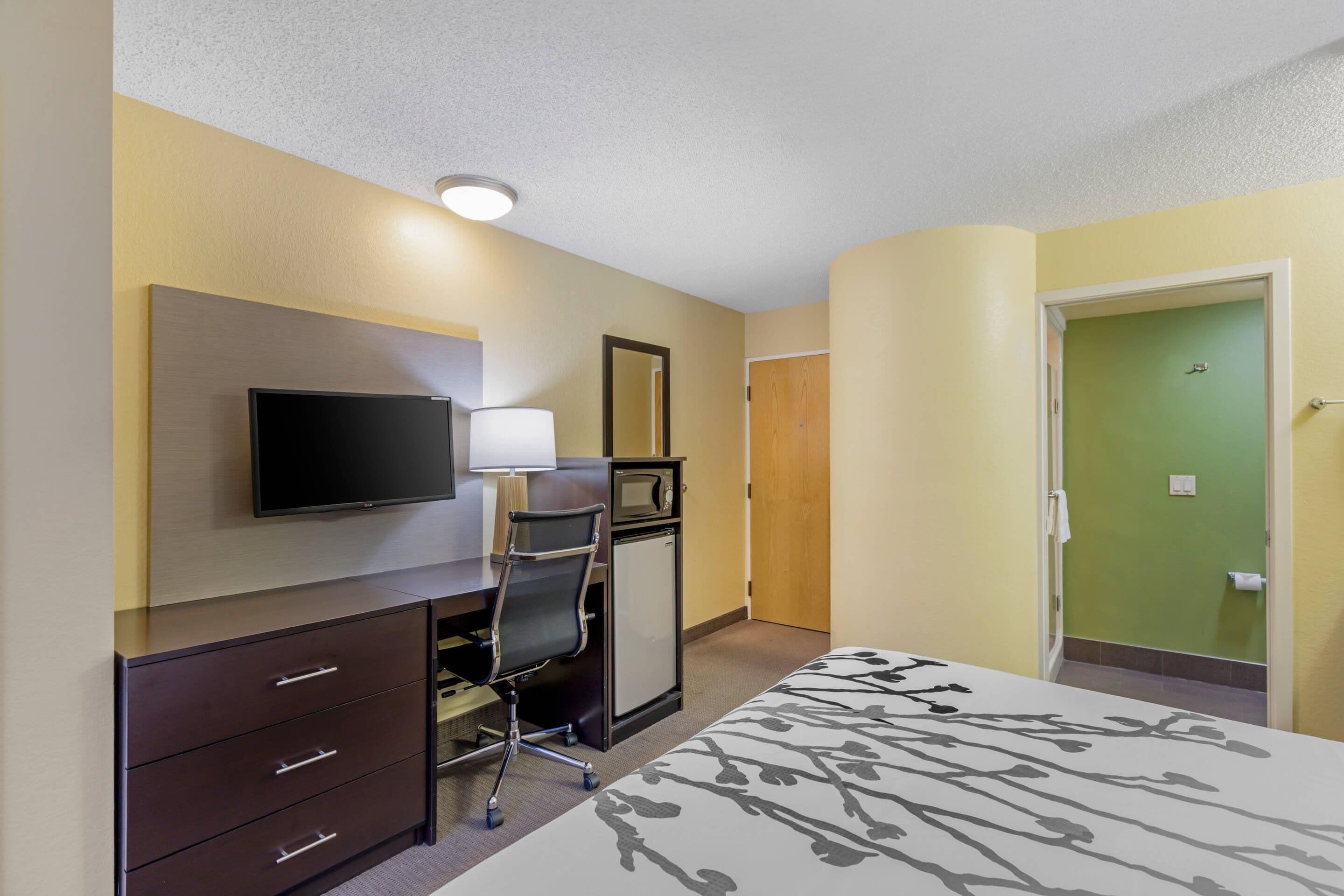 A hotel room with a bed, a desk with a chair, a television, a small cabinet, and a door leading to the bathroom.