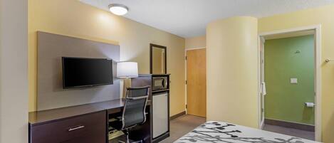 A hotel room with a bed, a desk with a chair, a television, a small cabinet, and a door leading to the bathroom.