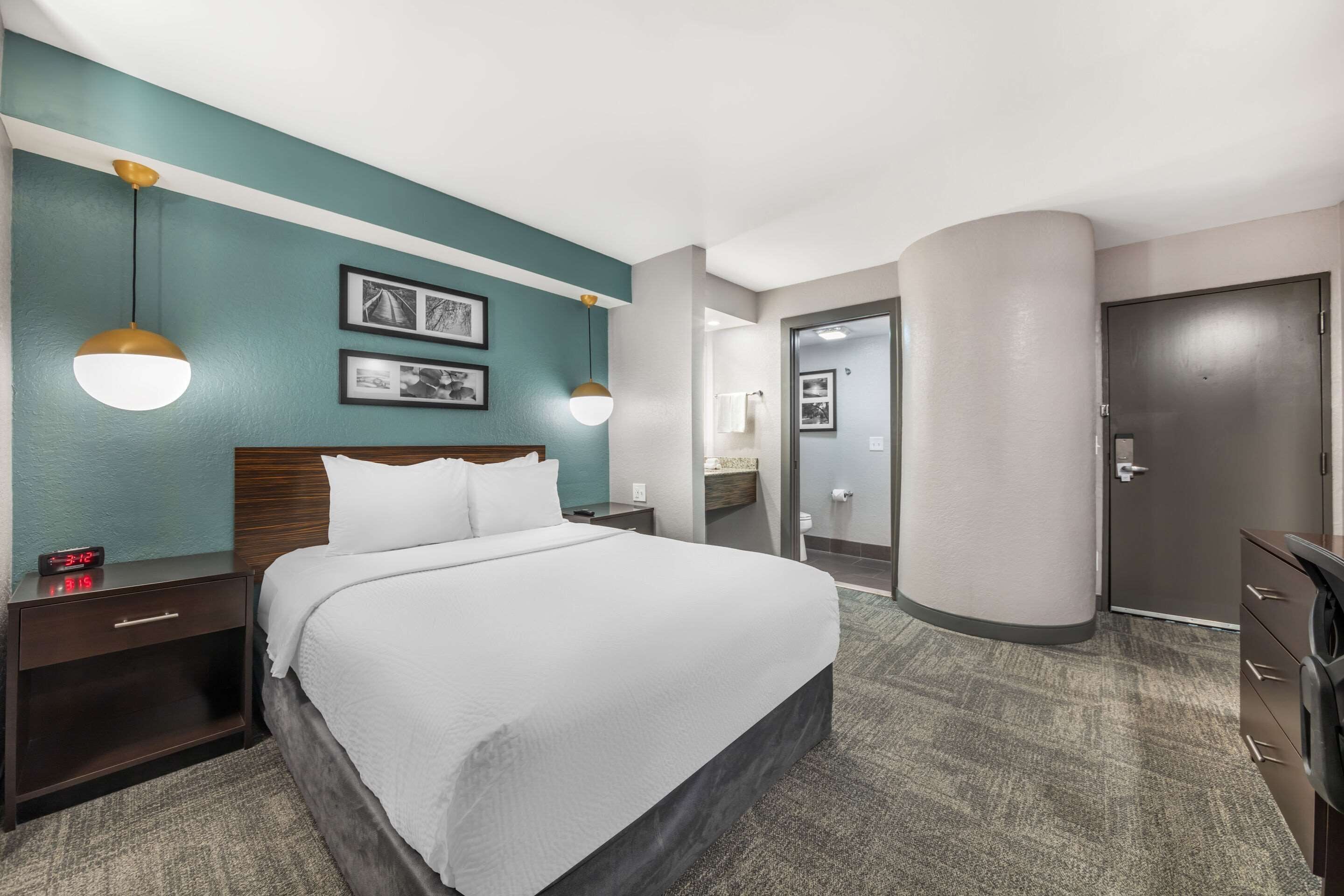 Habitación de hotel con cama, mesitas de noche, armario y baño visible a través de una puerta abierta.