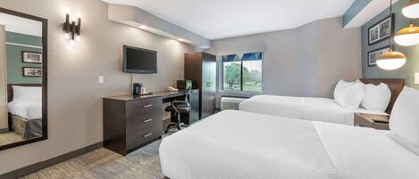 A hotel room with two beds, a desk with a computer, a TV, and a large window with a view of trees.