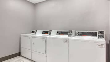 A row of four front-loading washing machines in a laundry room with a light-colored floor and walls.