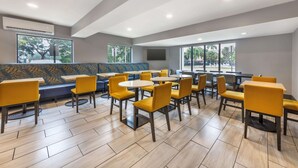 Un espace repas moderne avec des chaises et des tables jaunes, un banc à motifs bleus et de grandes fenêtres offrant une vue sur les arbres et un parking.