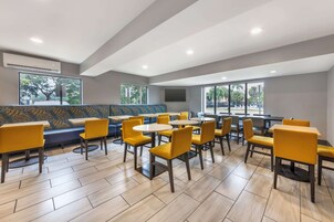 Un área de comedor moderna con sillas y mesas amarillas, un banco azul estampado, y ventanas amplias con vistas a árboles y un estacionamiento.
