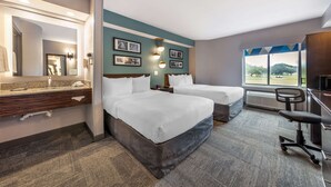 Une chambre d’hôtel avec deux lits, un bureau, une chaise, une grande fenêtre donnant sur un beau paysage, et une salle de bain avec un miroir et un lavabo.