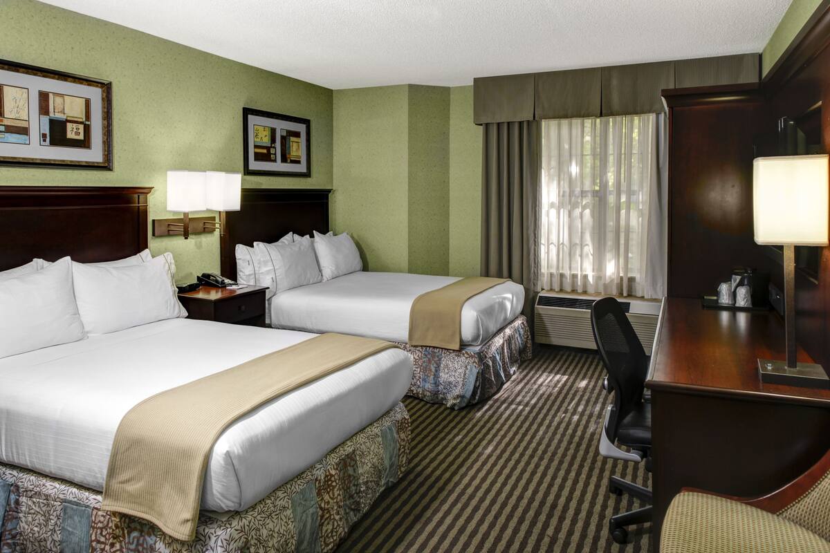 A hotel room with two beds, a desk, a chair, a lamp, and a window with curtains.