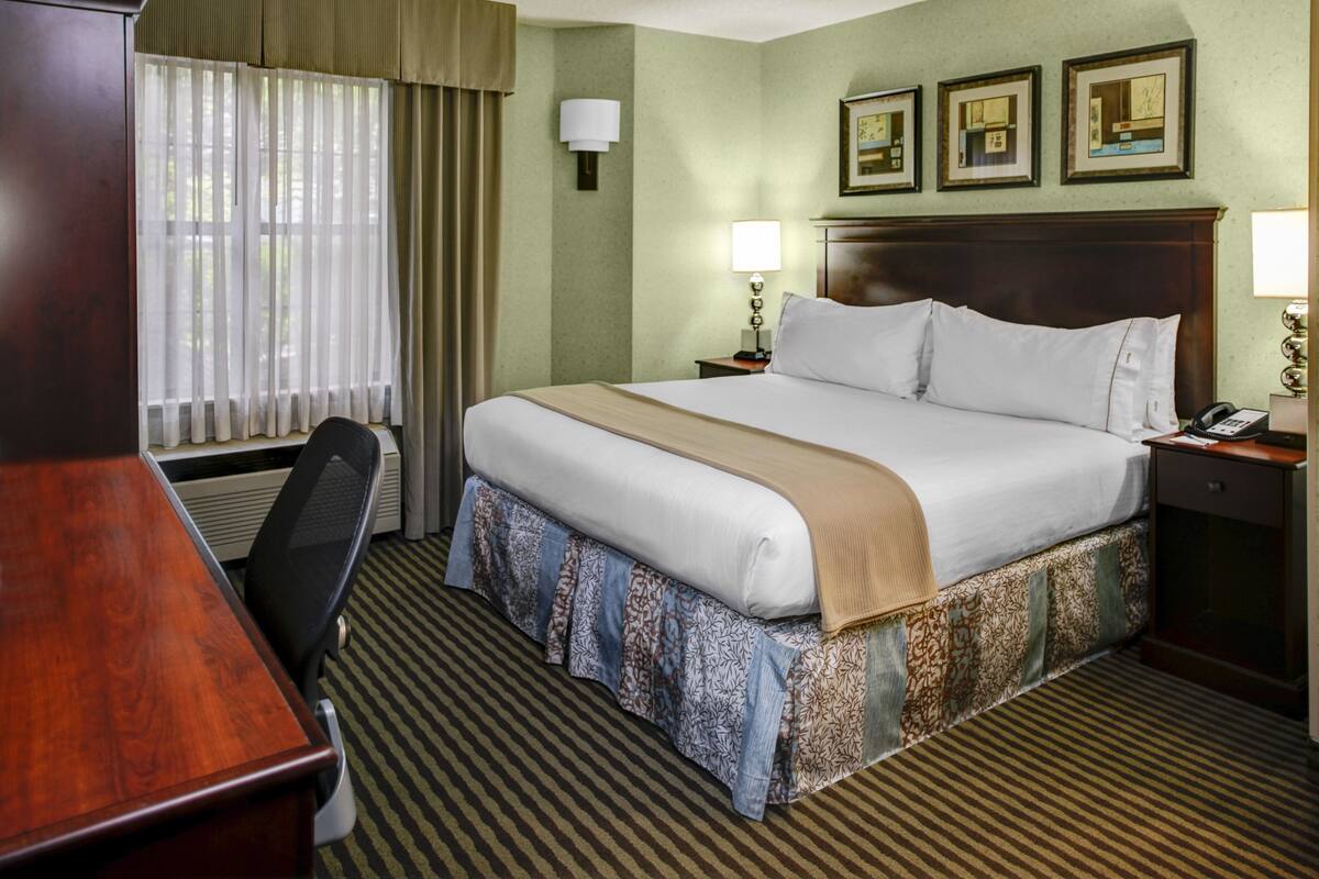 A hotel room with a large bed, a desk, and a chair. There are two bedside tables with lamps. The room has a window with curtains and a wall with framed pictures.