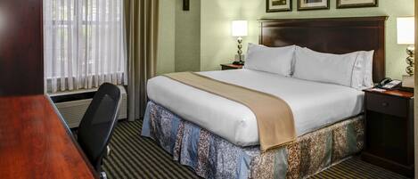 Una habitación de hotel con una cama grande, un escritorio y una silla. Hay dos mesitas de noche con lámparas. La habitación tiene una ventana con cortinas y una pared con cuadros.
