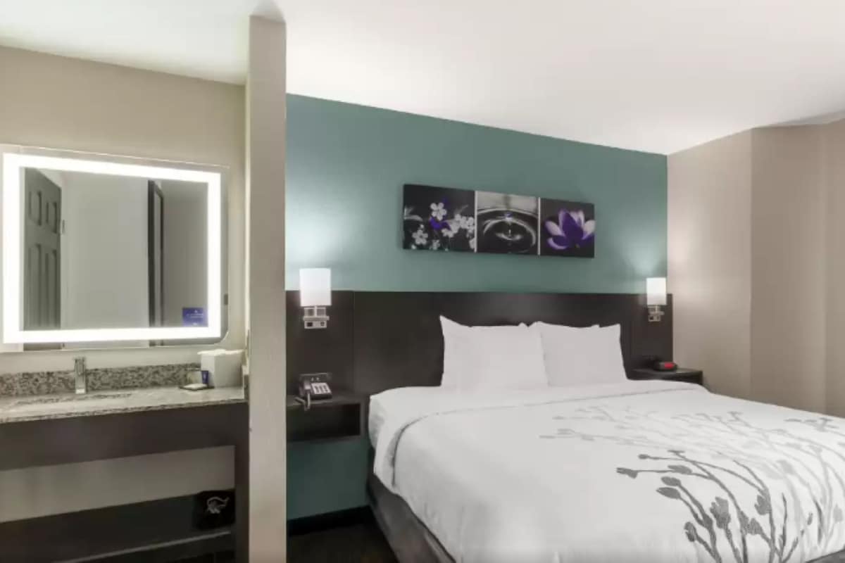 A hotel room with a bed, a large mirror, a vanity with a sink, and a wall-mounted artwork.