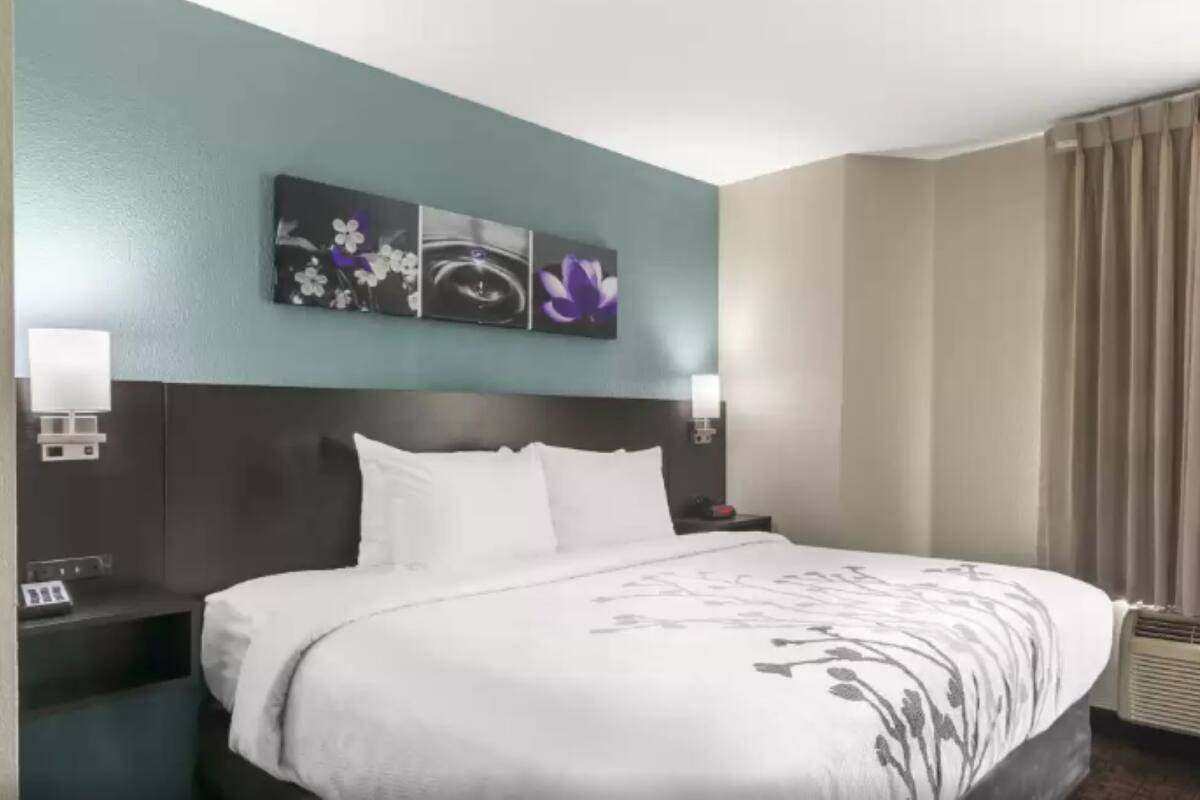 A hotel room with a large bed, a headboard with a clock, a wall-mounted artwork, a window with curtains, and a bedside table with a lamp.