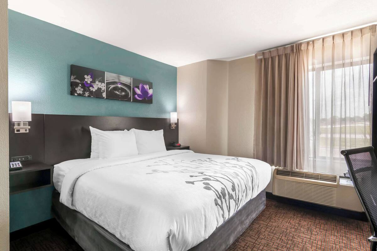 A hotel room with a bed, nightstands, a window with curtains, and a wall with floral artwork.
