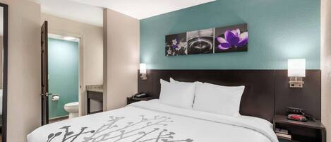 Habitación de hotel con una cama grande, una mesita de noche con lámpara, una obra de arte en la pared y un baño visible a través de una puerta abierta.