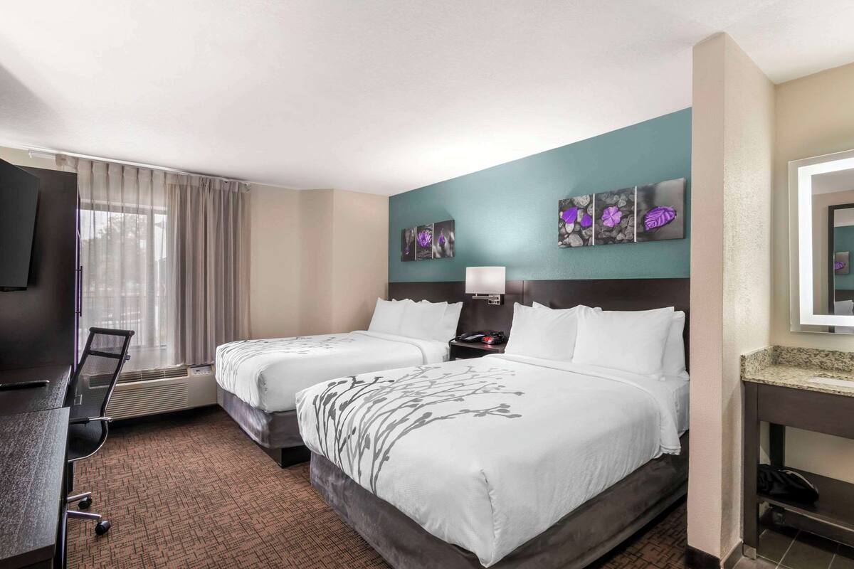 A hotel room with two beds, a desk, a chair, a TV, and artwork on the wall.