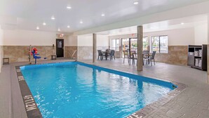 An indoor swimming pool with clear water, surrounded by stone walls and tiled flooring. There are chairs and tables in the background, and an exit sign is visible.
