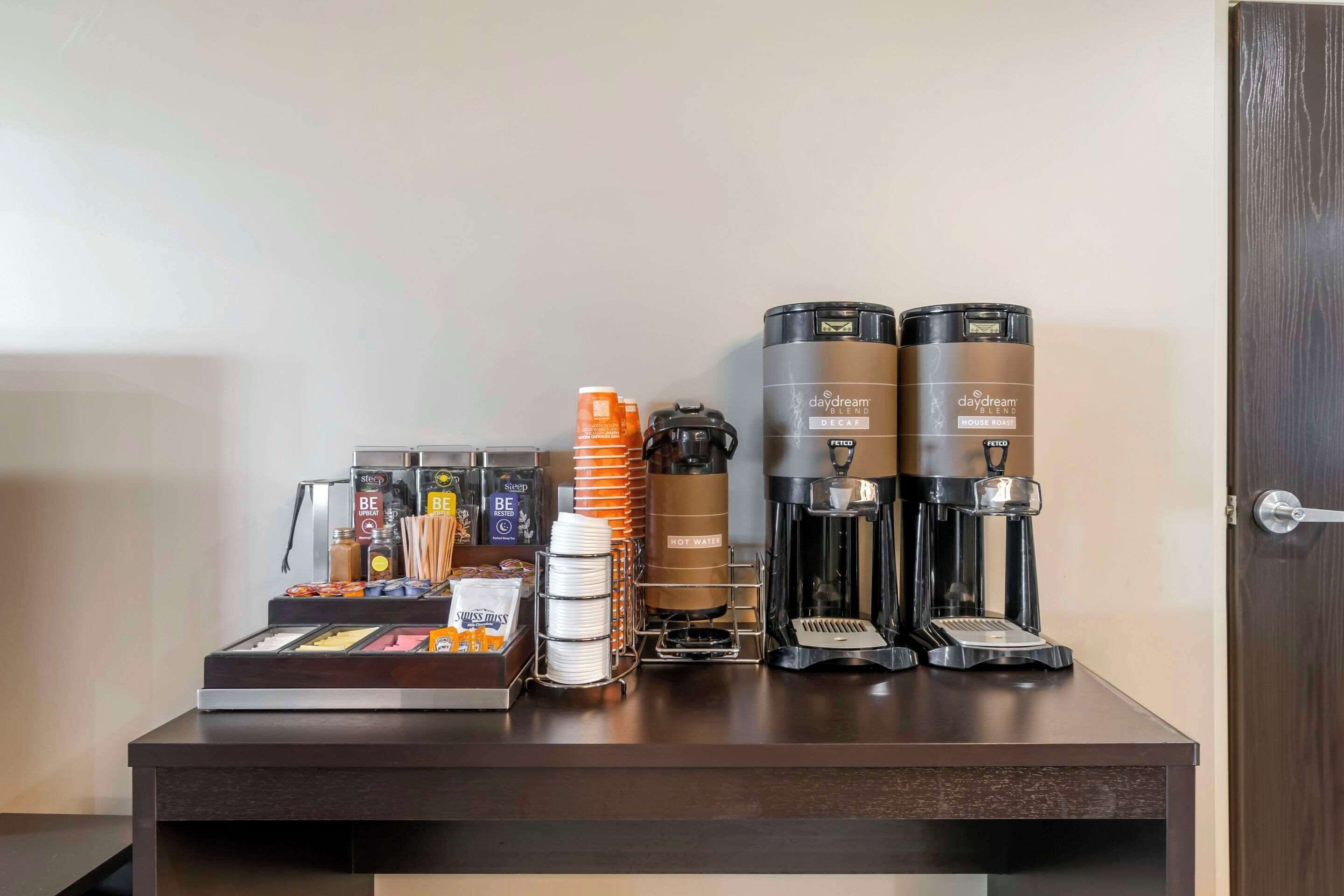 Una estación de café con dos cafeteras, un dispensador de agua caliente y diferentes cápsulas de café y utensilios.