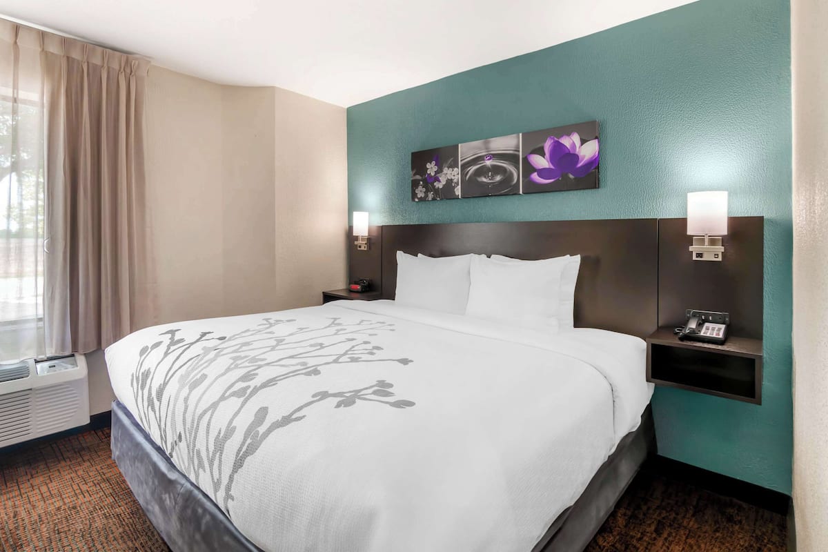 A hotel room with a large bed, a nightstand with a phone, a wall-mounted light, and a window with curtains.