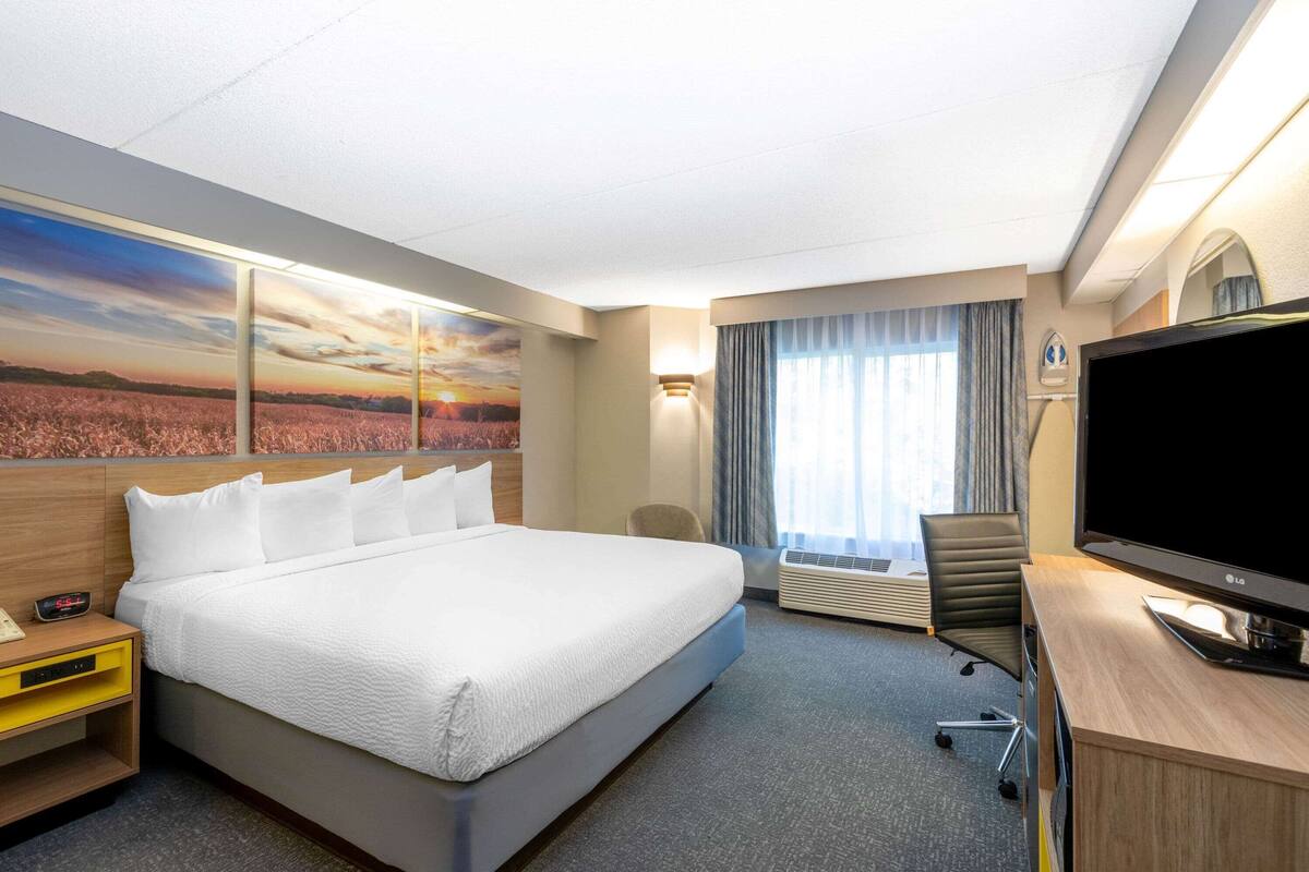 Una habitación de hotel con una cama grande, un escritorio con televisor, una silla, un equipo de aire acondicionado y un cuadro de un atardecer sobre un campo.