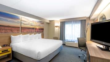 Una habitación de hotel con una cama grande, un escritorio con televisor, una silla, un equipo de aire acondicionado y un cuadro de un atardecer sobre un campo.