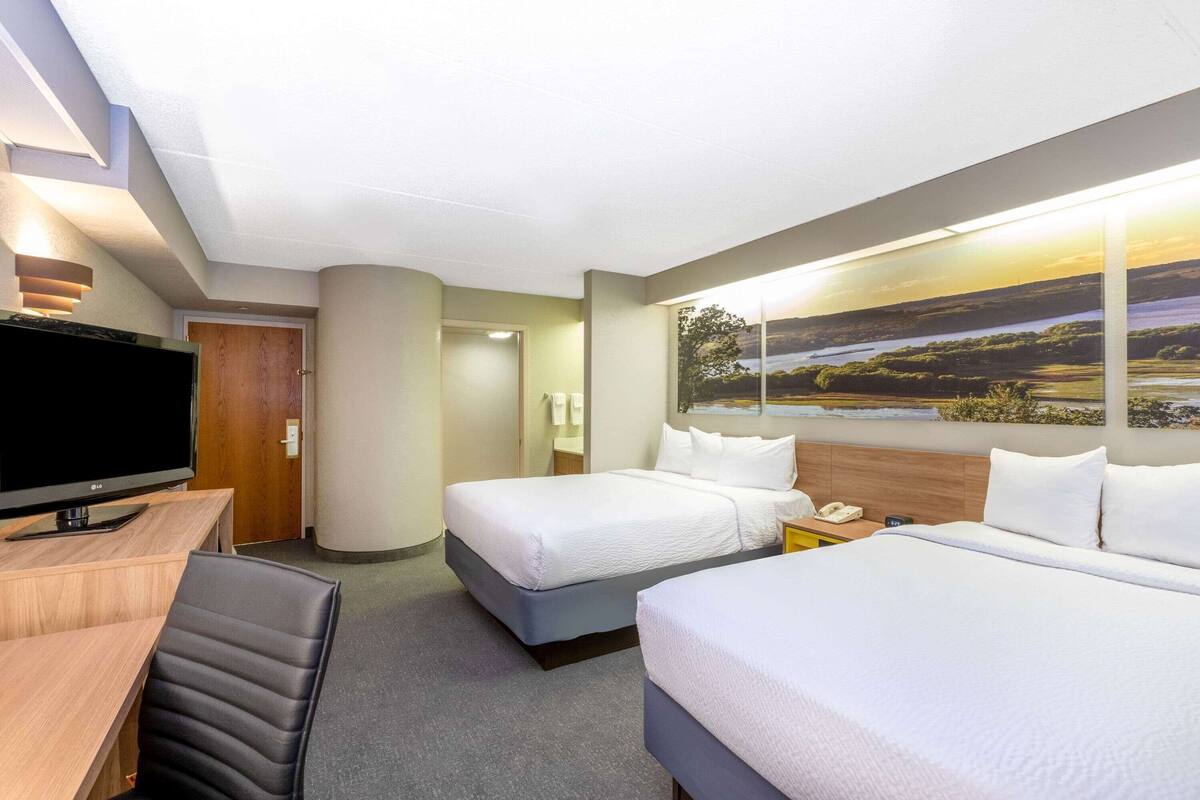 Habitación de hotel con dos camas, un escritorio con televisor y un ventanal con vistas al paisaje.