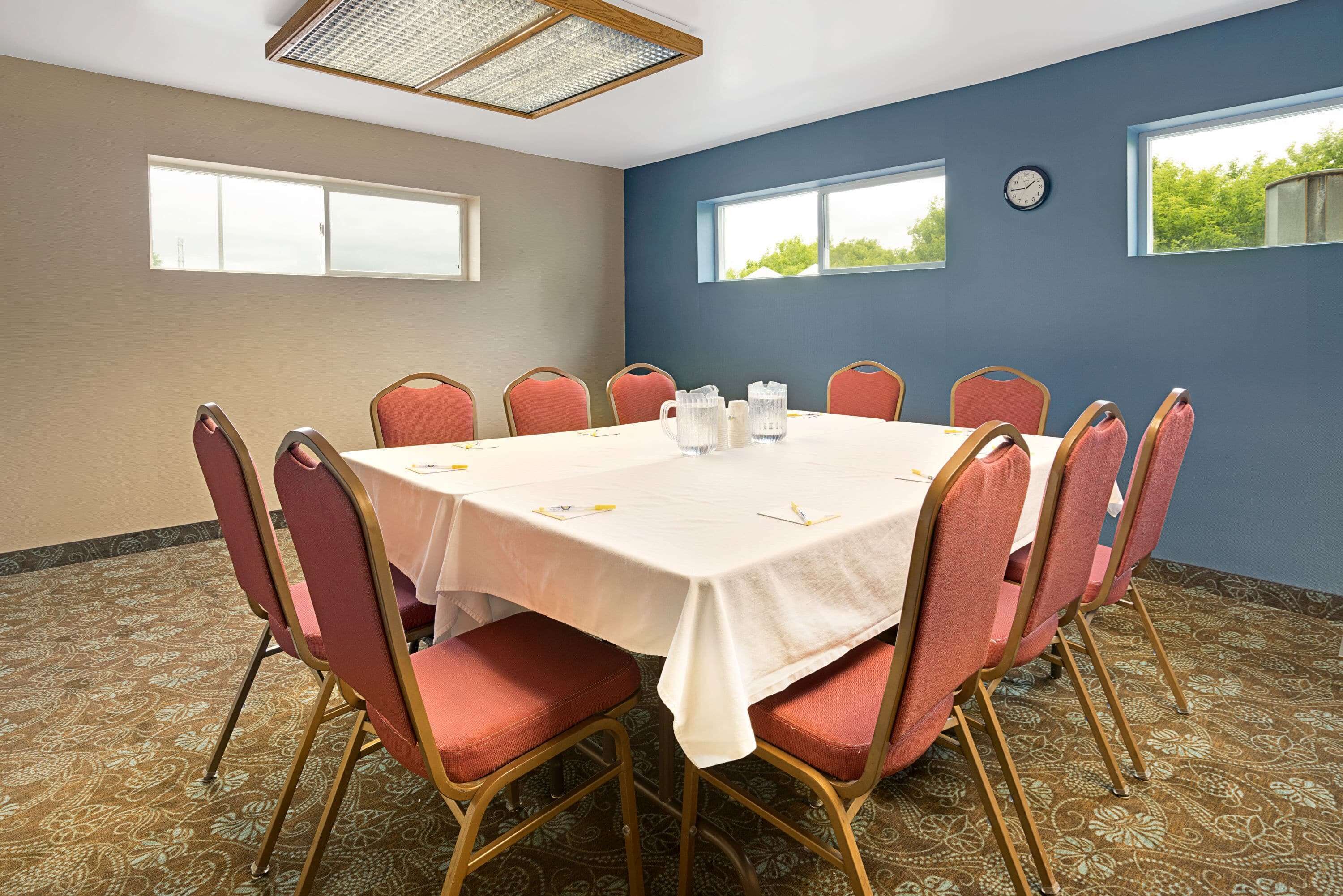 A conference room with a round table, red chairs, a clock on the wall, and windows with a view of greenery.