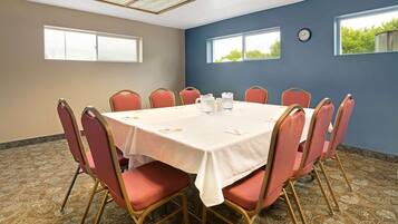 A conference room with a round table, red chairs, a clock on the wall, and windows with a view of greenery.