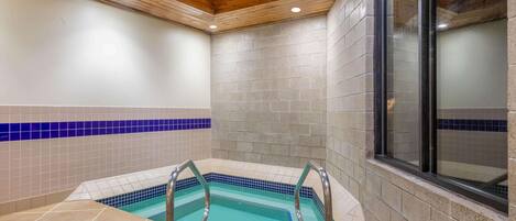 A swimming pool with a tiled border and metal ladder, surrounded by beige and blue tiles, with wooden ceiling beams and a brick wall.