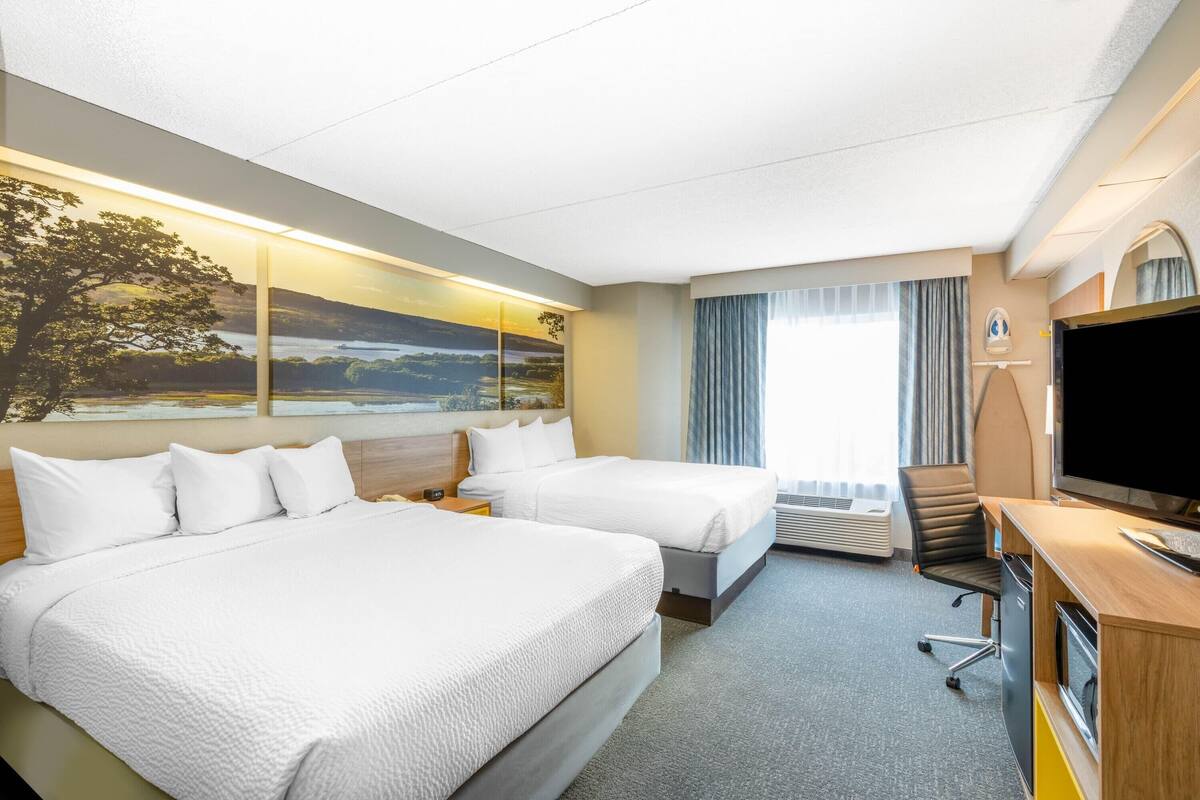Habitación de hotel con dos camas, un escritorio, un televisor de pantalla plana, un amplio ventanal con cortinas y un cuadro de un paisaje.
