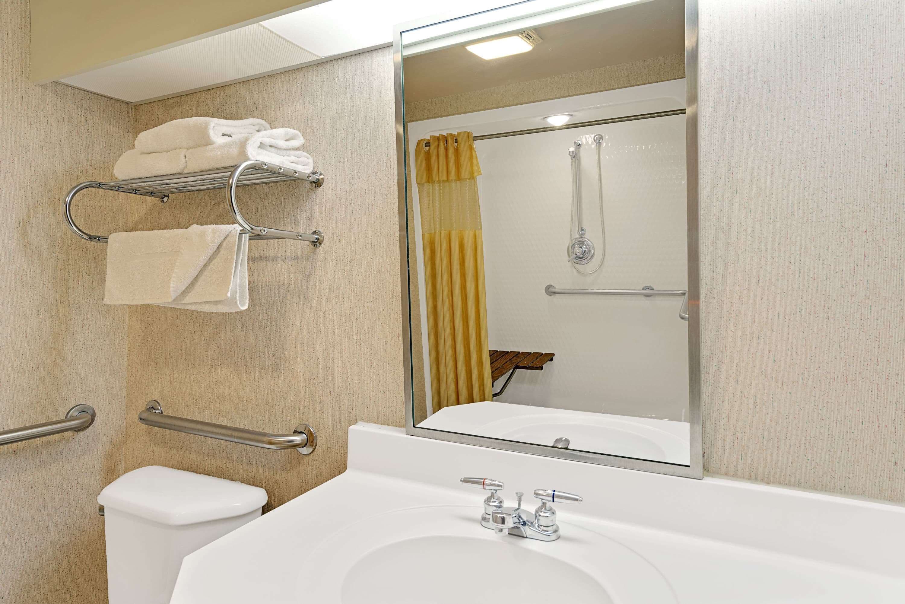 Un baño con un lavamanos blanco, un inodoro, un perchero con toallas dobladas, una ducha con cortina amarilla y un espejo que refleja el interior de la ducha.