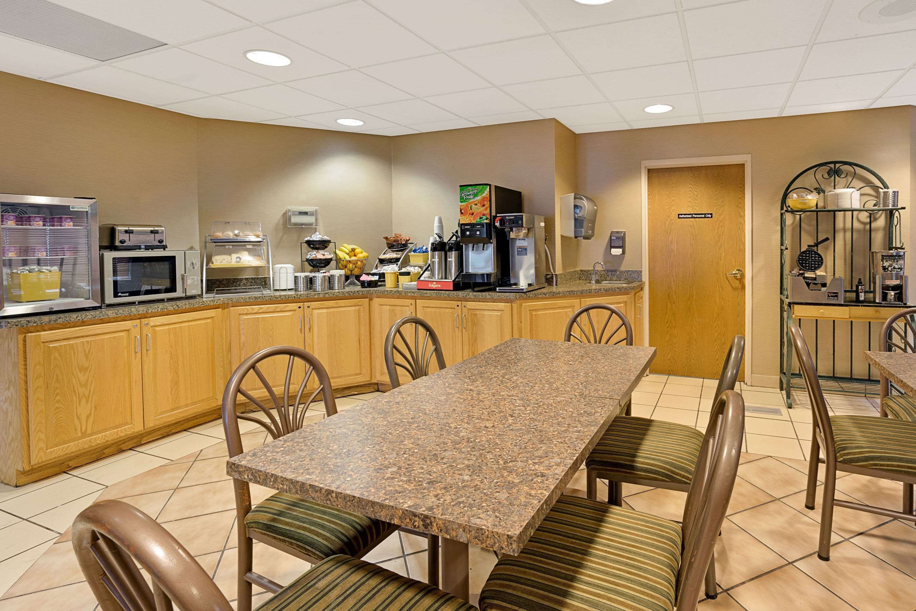 A hotel breakfast area with a long table, chairs, a microwave, coffee maker, and a fruit display.