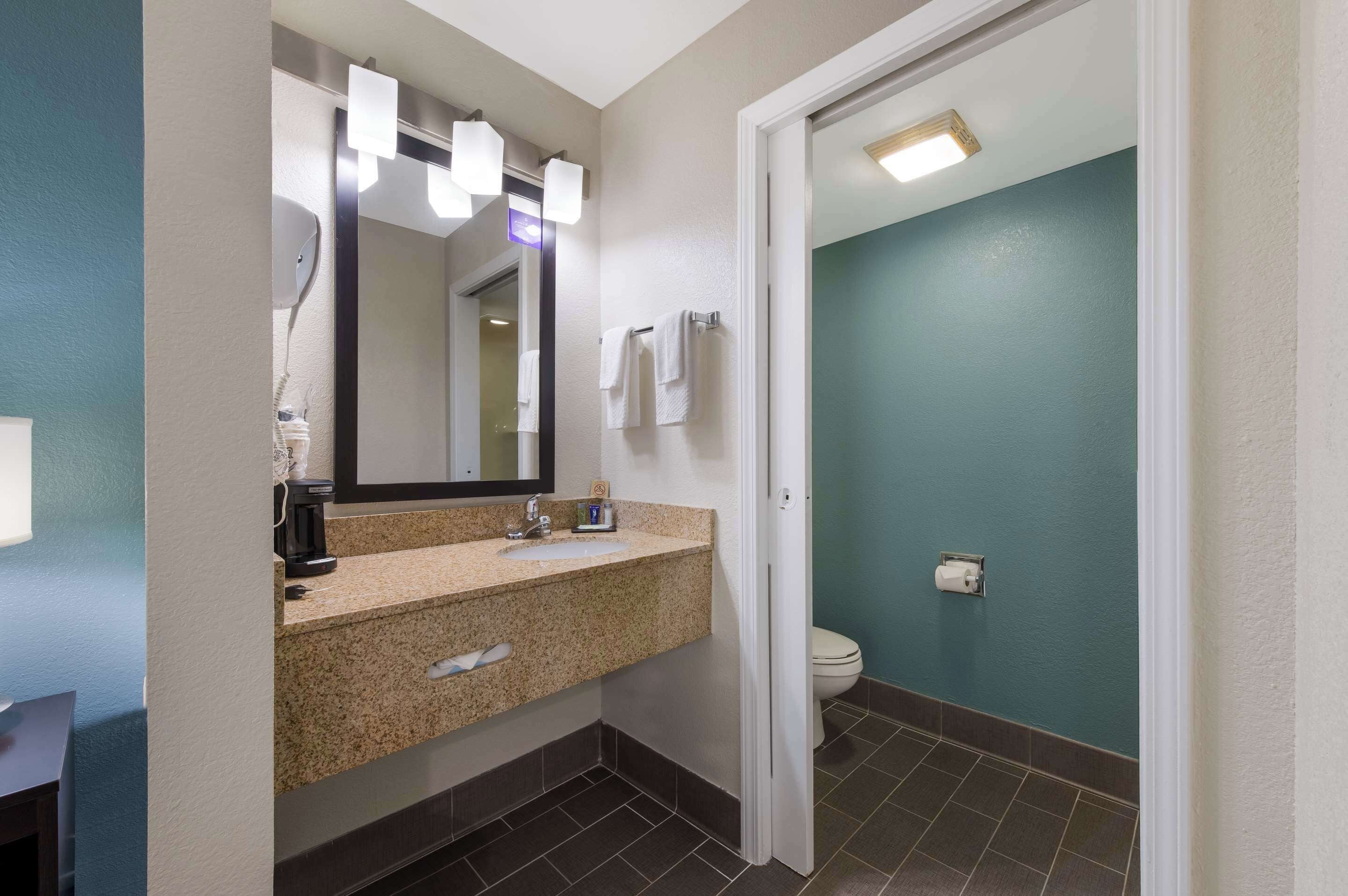 Une salle de bain avec un lavabo surmonté d’un plan de travail en granit, un miroir, une machine à café et des toilettes.