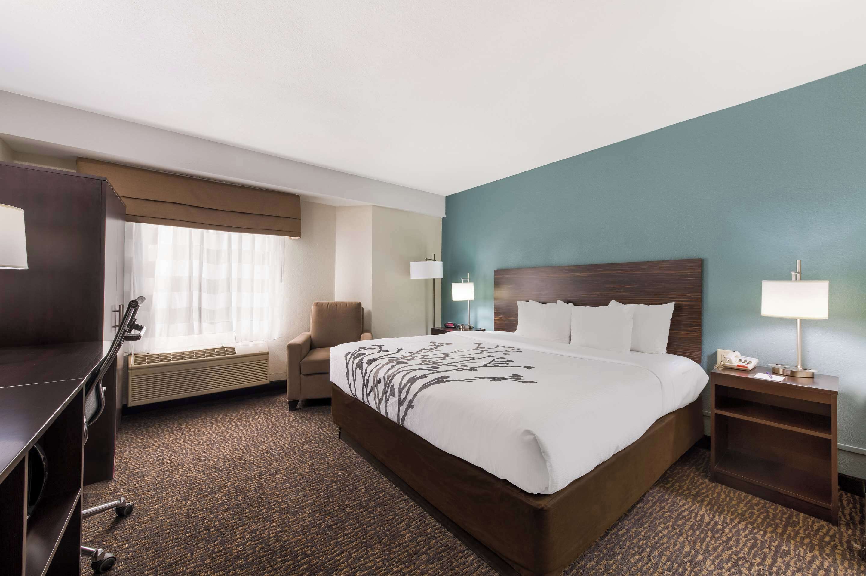 A hotel room with a large bed, a desk, a chair, a desk lamp, and a nightstand.