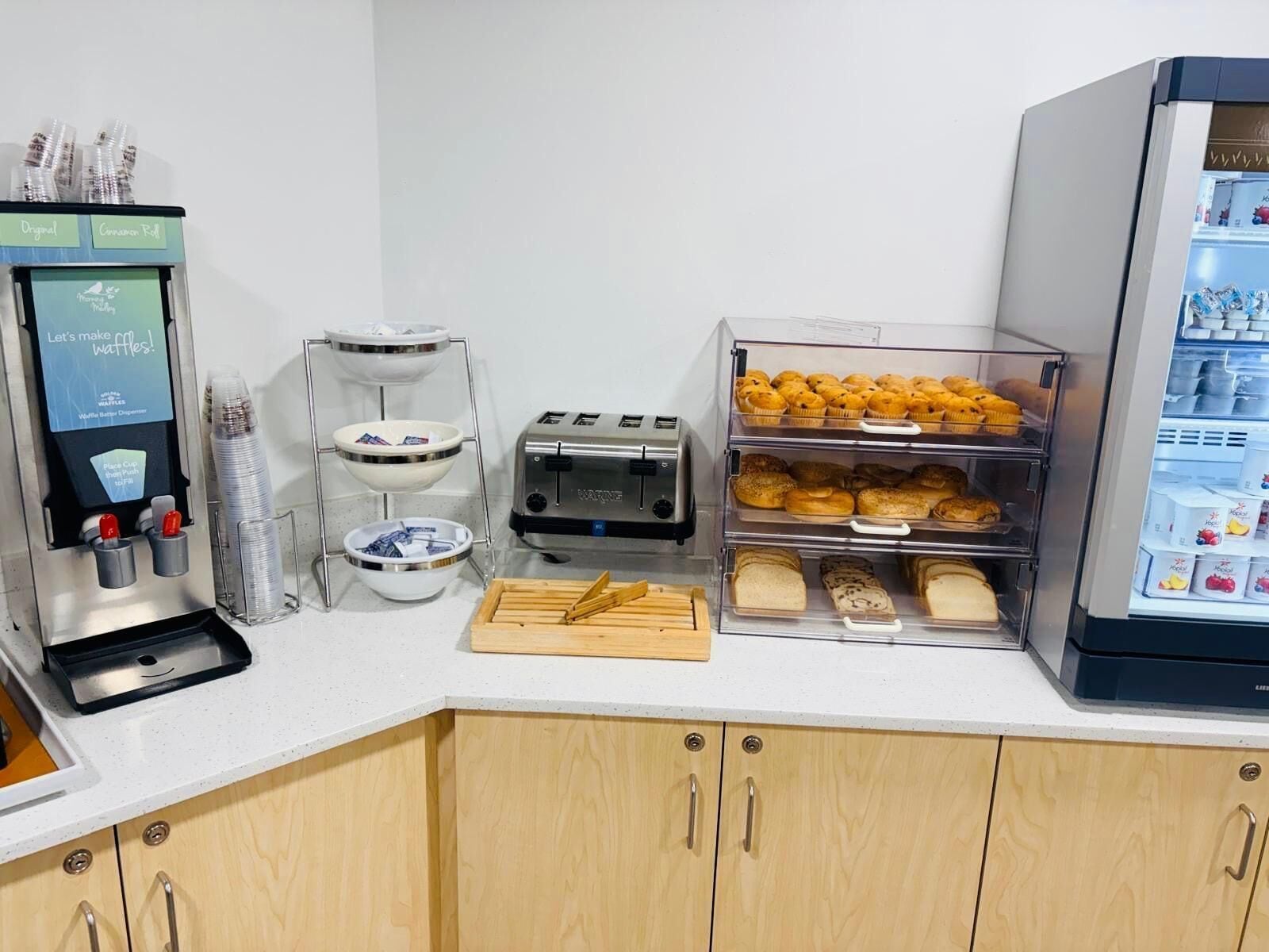 A kitchenette with a waffle maker, toaster, bread, and a refrigerator stocked with beverages.