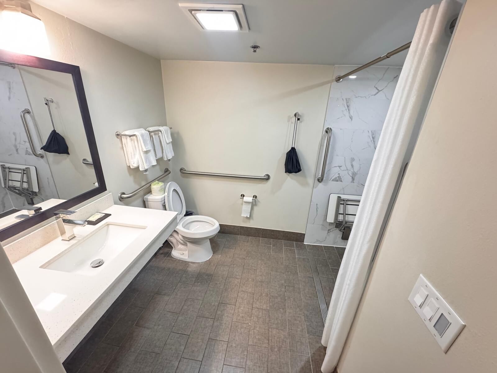 Et badeværelse med håndvask, toilet, spejl, håndklædestang, gelænder og en bruseplads.