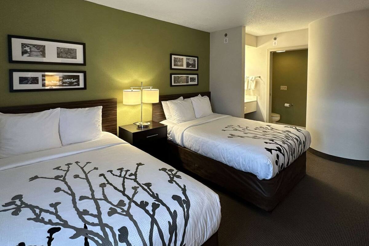 A hotel room with two beds, a nightstand with a lamp, framed pictures on the wall, and a bathroom visible through an open door.