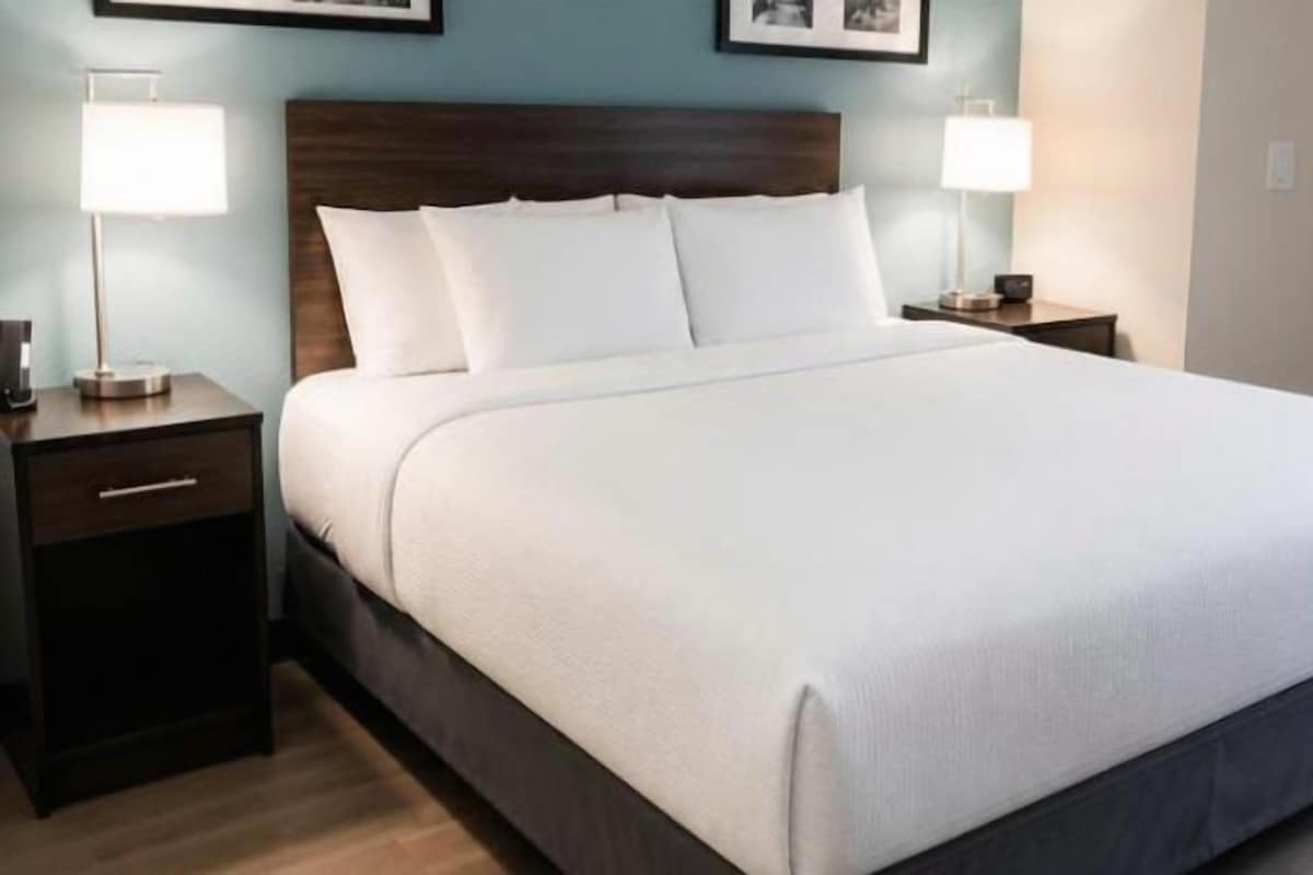 Una cama grande con cabecera de madera, dos mesitas de noche con lámparas, y una ventana con cortinas.