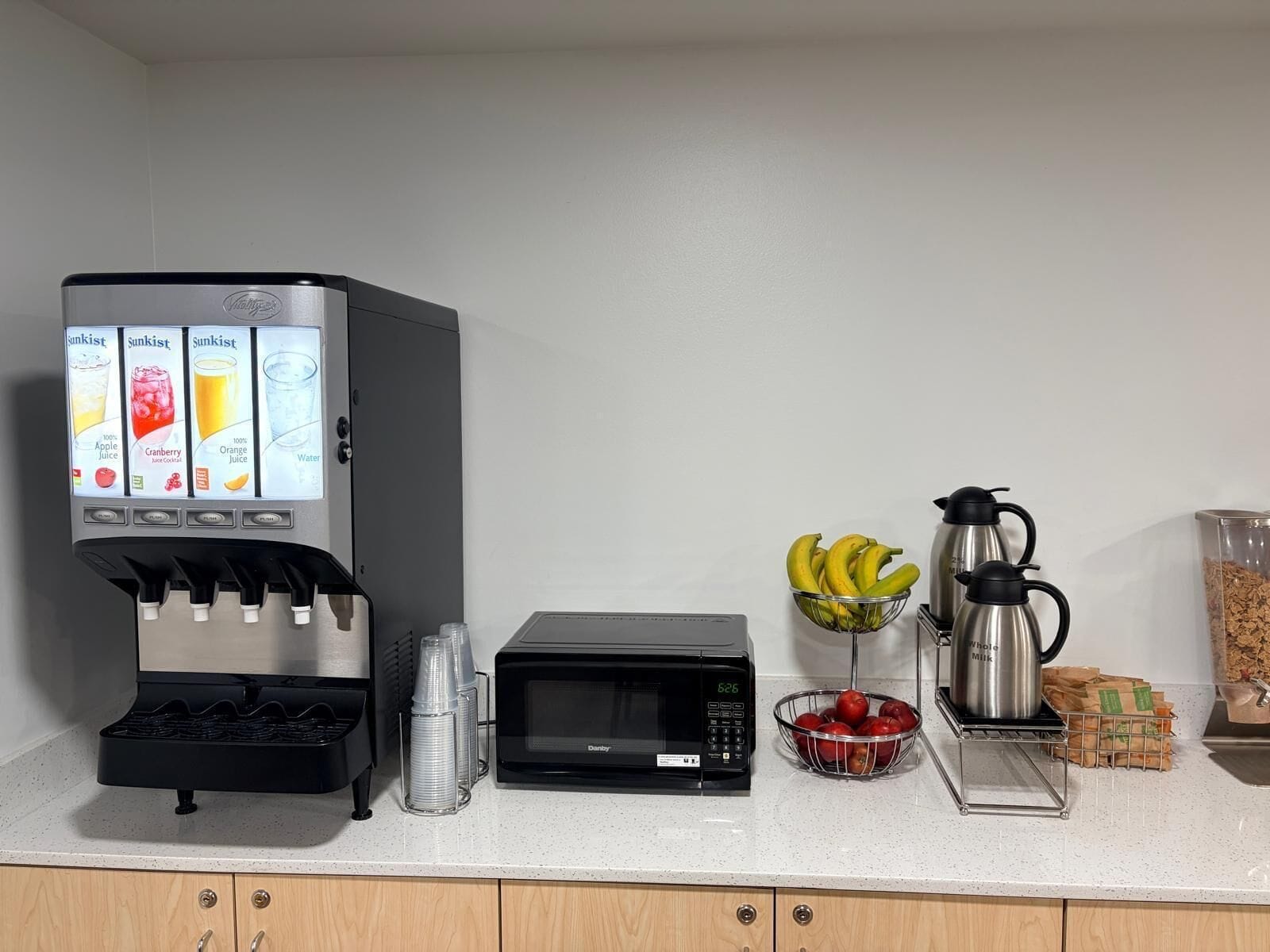 Una encimera de cocina con un dispensador de jugo, un microondas, un cuenco de plátanos, un cuenco de manzanas, una cafetera y un recipiente con cereal.