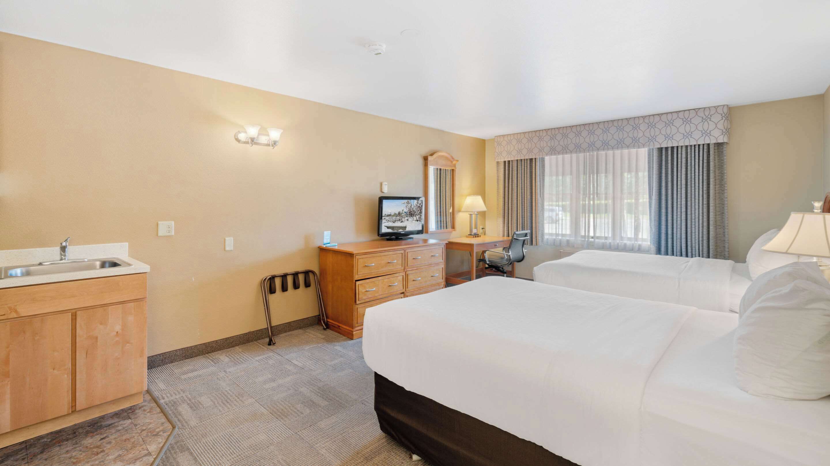 A hotel room with two beds, a sink, a mirror, a television, and a desk with a lamp.