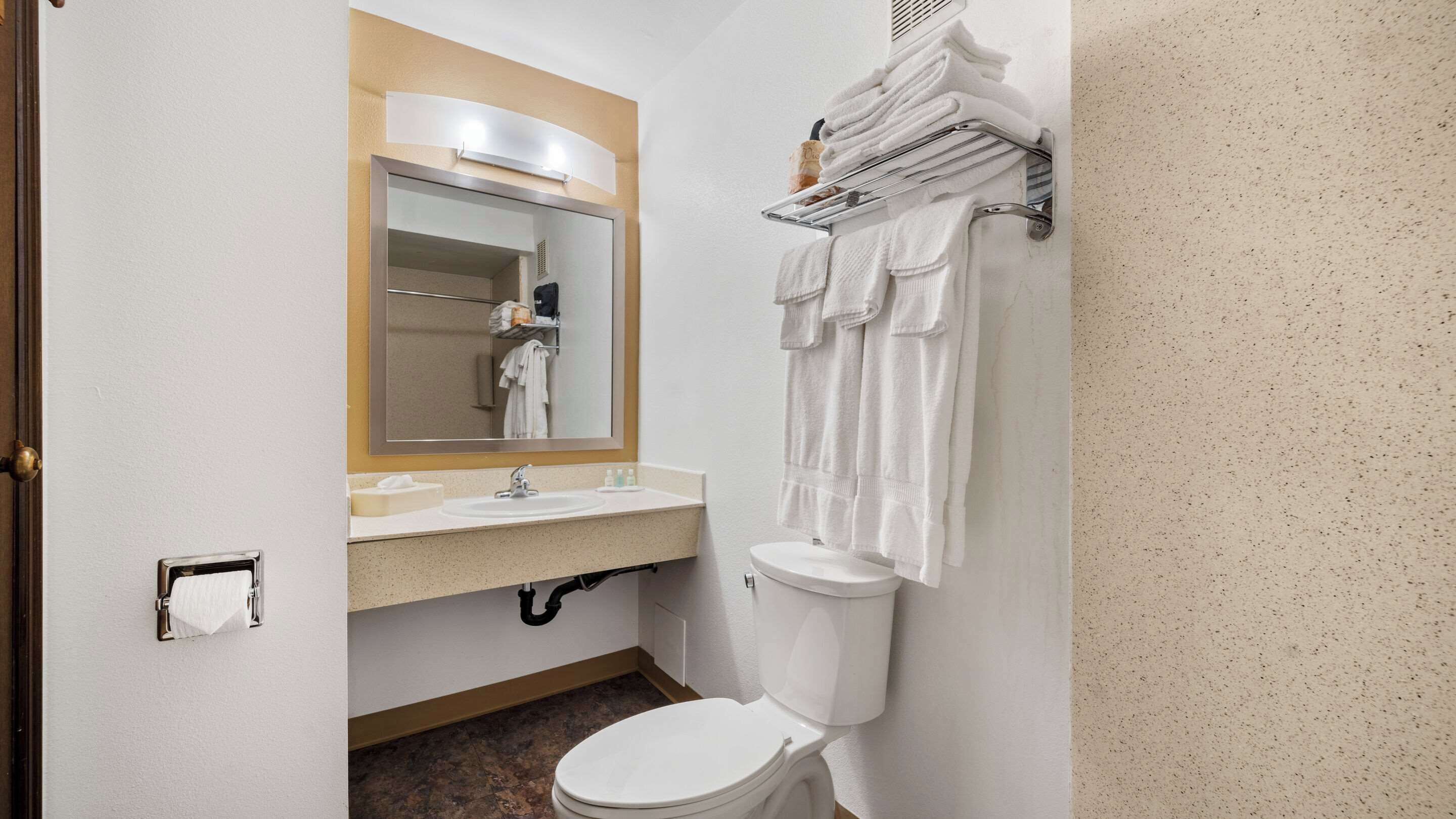 Un baño con lavamanos, espejo e inodoro. Hay un perchero con toallas y un pequeño estante con artículos de aseo.