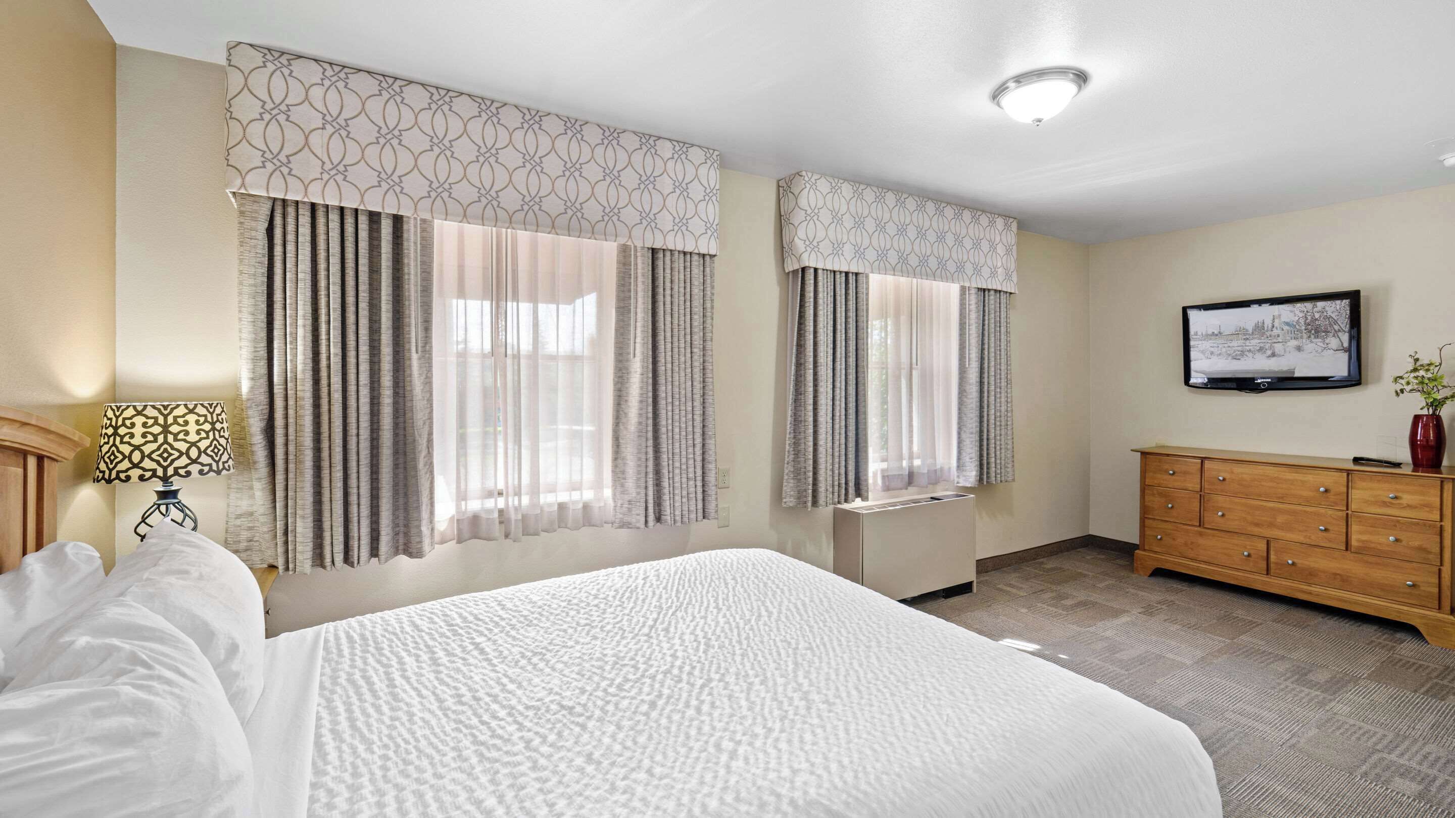 A hotel room with a large bed, two windows with curtains, a wooden dresser, a TV, and a lamp.