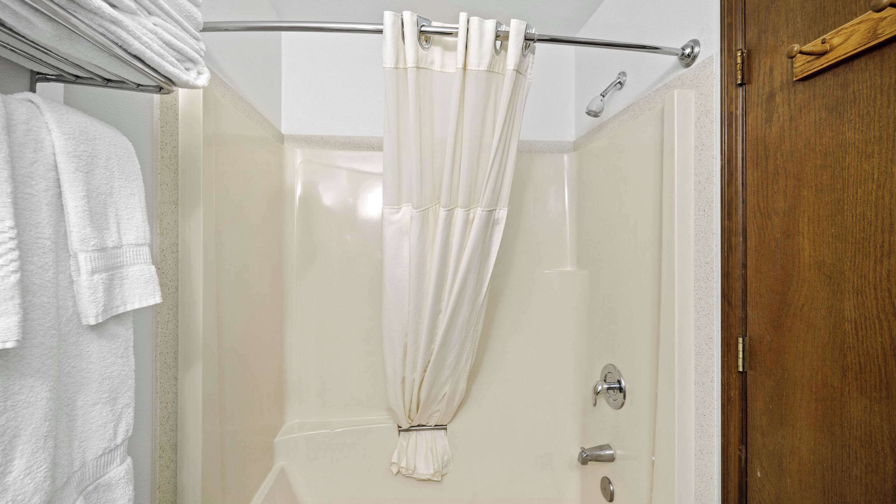 A shower with a white curtain, a towel rack with white towels, and a wooden door.