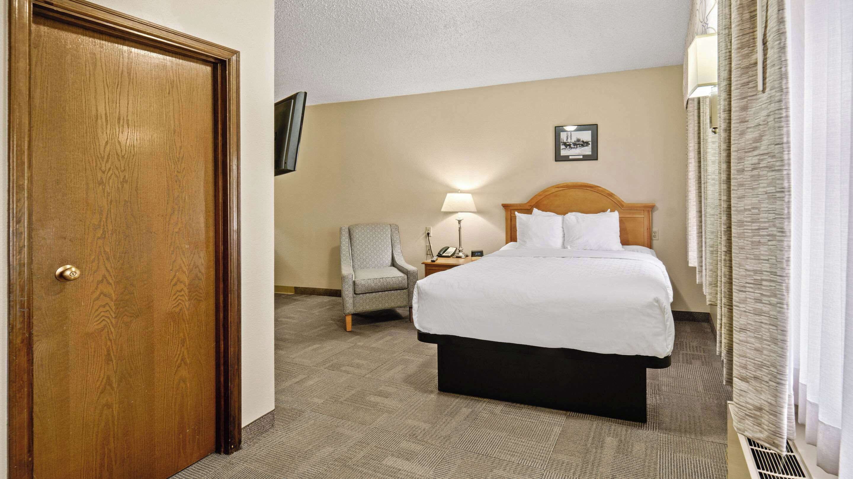 A hotel room with a wooden door, a bed with white bedding, a chair, a nightstand with a lamp, and a framed picture on the wall.