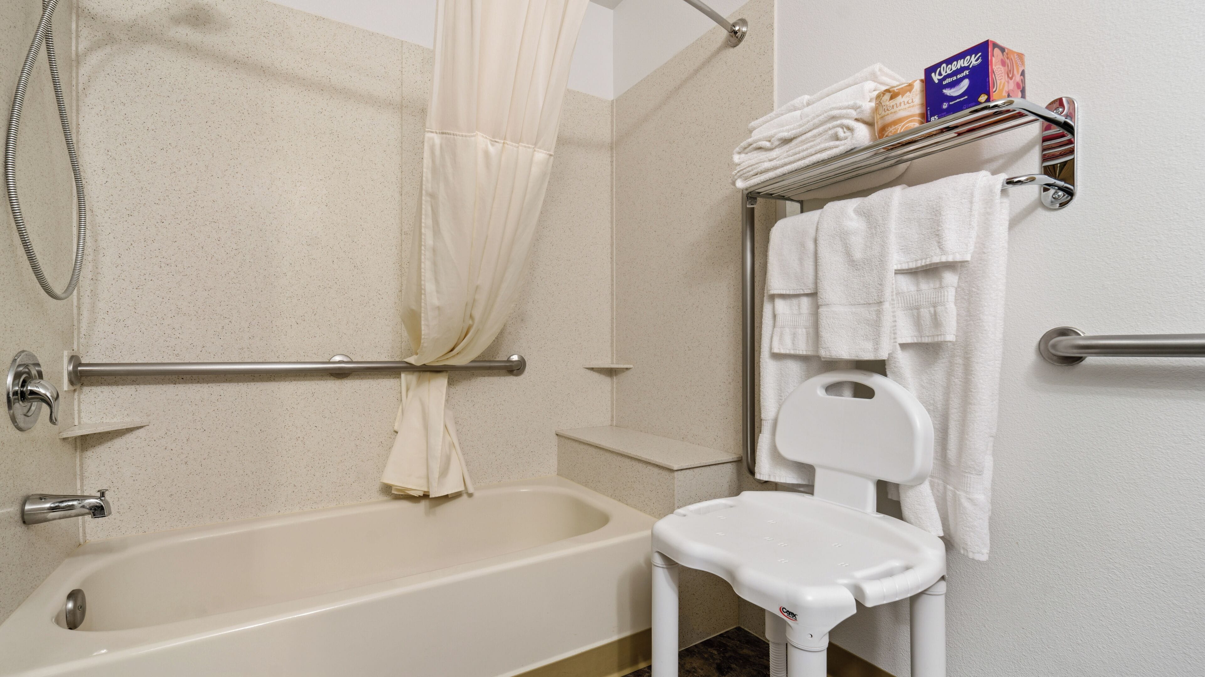 Un baño con bañera, cortina de ducha, una silla blanca, toallero con toallas dobladas y una caja de Kleenex.
